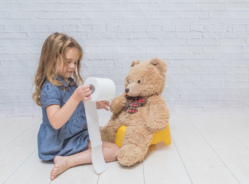 Young girl holding toilet paper while potty training a teddy bear on a training potty.